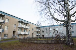 Ett hyreshus i tre våningar med beiga väggar och blå accenter visas. I förgrunden finns en liten inhägnad lekplats med rutschkana och gungor. Ett kalt träd står i närheten, och cyklar står parkerade vid byggnaden, som förvaltas av Överkalixbostäder för hyreslägenhetsentusiaster.