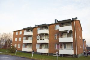 Hyreshus i tre våningar med flera balkonger och stora fönster, förvaltas av Överkalixbostäder. Strukturen har ett platt tak och är omgiven av en gräsyta. Några träd syns i bakgrunden under en halvmolnig himmel, typiskt för hyreslägenheter.