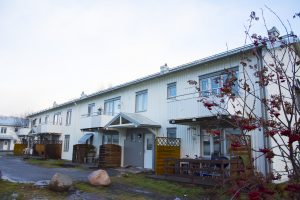 En rad moderna, vita radhus, som liknar stilfulla hyreslägenheter, ståtar med små balkonger och verandor. Himlen är mulen, med buskar av röda bär i förgrunden. En gångväg och stora stenar leder till dessa inbjudande hem.