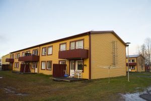 En tvåvånings gul byggnad med röda accenter erbjuder charmiga balkonger och en mysig veranda. Överkalixbostäder presenterar denna hyreslägenheter med gräsbevuxen gård, klädstreck och snöfläckar. Den klara himlen antyder en strålande solig dag.