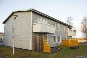 Ett grått trähus i två våningar med två balkonger på övre plan framkallar charmen hos en Överkalixbostäderfastighet. Den nedre våningen har trästaket och små trädgårdar. En gatlykta står i närheten, och bladlösa träd syns mot den klara himlen, vilket förstärker dess fridfulla atmosfär.