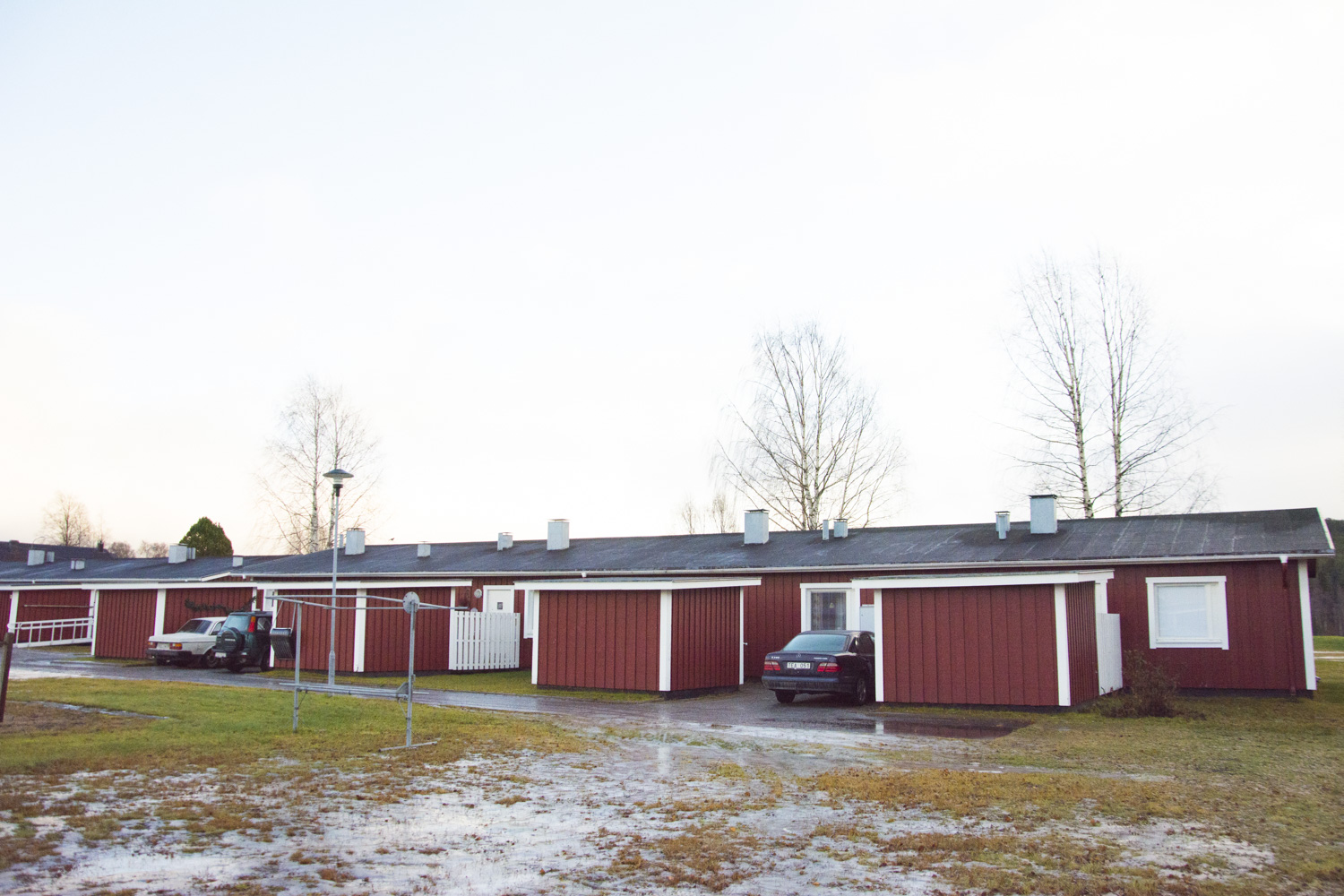 Overkalixbostader6 En rad rödvita enplanshus med platta tak en molnig dag som påminner om Överkalixbostäders design. Två bilar står parkerade på uppfarter, medan snöfläckar prickar gräset. Bladlösa träd står i bakgrunden och fullbordar denna fridfulla bild av svenska hyreslägenheters boende.