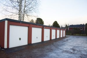 En rad med röda och vita garageportar, var och en stängd, kantar en asfalterad yta, vilket speglar Överkalixbostäders prydliga estetik. Ett kalt träd står i slutet med bostadshus och buskar i bakgrunden under en klar himmel.