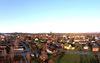 Flygfoto över en liten stad med färgglada hus och byggnader under en klarblå himmel. En kyrka med högt torn är framträdande i centrum, medan hyreslägenheter inbäddade bland träd och öppna fält lyfter fram den lantliga charmen.