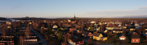Flygfoto över en pittoresk by vid soluppgången, med färgglada trähus och en framstående kyrkspira i mitten. Landskapet, som påminner om Överkalixbostäders design, har träd, vägar och avlägsna kullar under en klar himmel.
