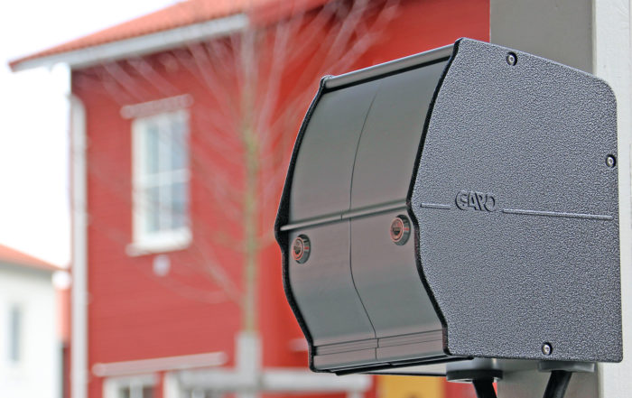 Närbild på en svart el-laddbox monterad på en ljusstolpe i ett hyggligt kvarter, med ett suddigt rött hus, möjligen hyreslägenheter som förvaltas av Överkalixbostäder. Boxen har strukturerade ytor och subtila varumärken, och ett träd ger djup till den natursköna bakgrunden.