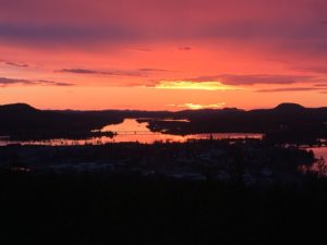 En fantastisk solnedgång över en landskapsvy med en flod som reflekterar himlens livliga färger. Horisonten har silhuettade kullar under en gradient av djupröda, orange och rosa nyanser. Spridda moln förstärker den dramatiska effekten och erbjuder lugn som liknar lugna stunder i Överkalixbostäder.