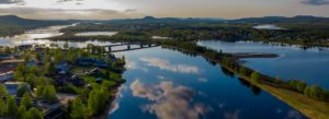 Flygfoto över ett fridfullt landskap med en bred flod som reflekterar himlen och molnen. Omgiven av frodig grönska prickar små hus och hyreslägenheter längs flodstränderna. Flera broar korsar vattnet, och avlägsna kullar är synliga under en delvis molnig himmel.