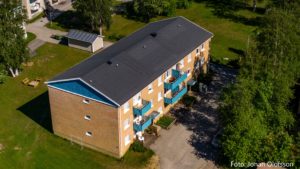 Flygfoto över ett rektangulärt lägenhetshus i två våningar i tegel med blå balkonger, inbäddat i frodig grönska. Ett skjul kikar fram bakom träden. Denna charmiga hyresfastighet förvaltas av Överkalixbostäder. Fotokrediten är synlig.