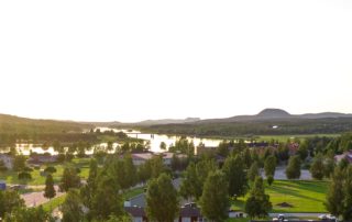 En naturskön flygvy över ett landsbygdslandskap vid solnedgången visar träd, en idrottsplats, hyreslägenheter och en flod. Böljande kullar och en ljus himmel ramar in scenen i Överkalixbostäder och skapar en lugn atmosfär.