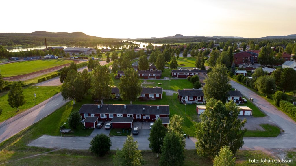 DJI_0412 text Flygfoto över ett villakvarter med rödvita byggnader, eventuellt Överkalixbostäder hyreslägenheter, omgiven av träd. En flod rinner i bakgrunden under en nedgående sol och kastar ett varmt sken på landskapet. Böljande kullar syns i fjärran.