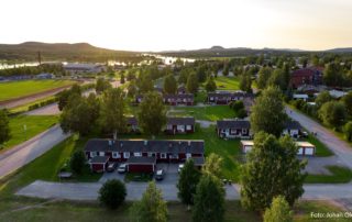 Flygfoto över ett villakvarter med rödvita byggnader, omgiven av träd. En älv rinner i bakgrunden under en nedgående sol och kastar ett varmt sken på landskapet. Böljande kullar syns i fjärran.