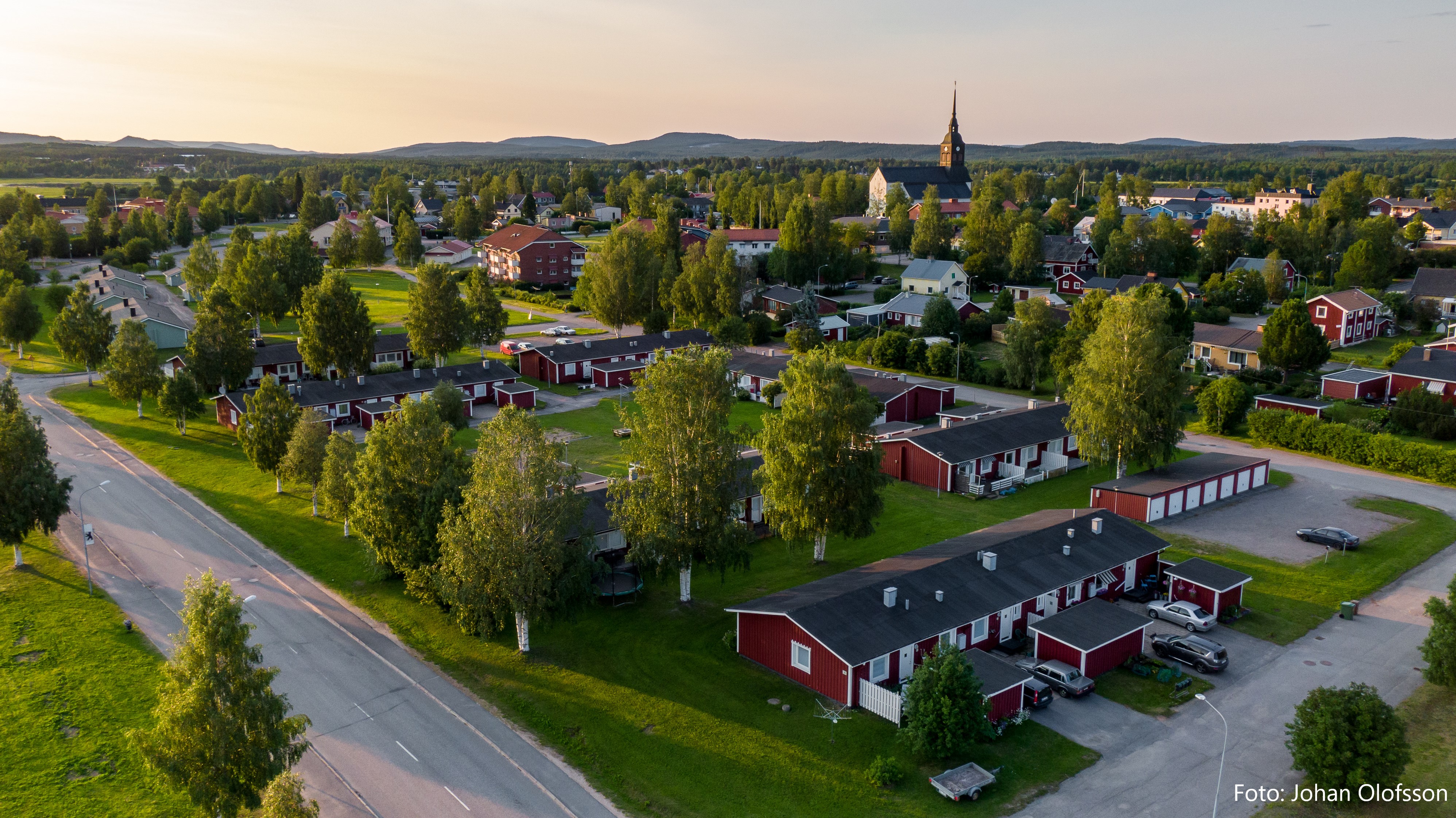 DJI_0414 Text Flygfoto över en liten stad som visar upp röda träbyggnader och frodiga träd med en kyrka i bakgrunden. Vägen kröker sig mjukt genom gröna gräsytor under klar himmel och solar sig i varmt ljus – en idyllisk scen som påminner om Överkalixbostäders charmiga hyreslägenheter.