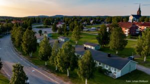 Flygfoto över ett förortskvarter med rader av hus med gröna tak, troligen en del av Överkalixbostäder, omgiven av träd och öppna fält. En kyrka syns i bakgrunden. Bilden är tagen under solnedgången och kastar varmt ljus över landskapet.