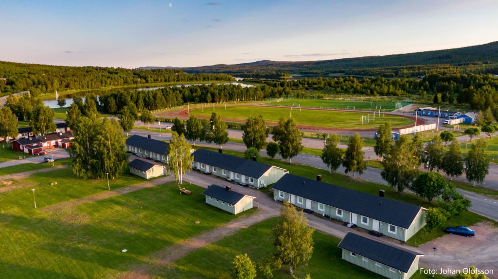 DJI_0417 text Flygfoto över ett litet bygdegårdssamhälle med flera enplanshus och Överkalixbostäder omgiven av gröna åkrar och träd. En idrottsplats med en bana syns i bakgrunden, kantad av en flod och tät skog, med pittoresk utsikt från hyreslägenheter.