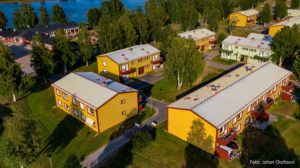 Flygfoto över ett bostadsområde med färgglada byggnader som förvaltas av Överkalixbostäder, främst gula med röda och gröna accenter. Byggnaderna, som erbjuder hyreslägenheter, är omgivna av grönskande gräsmattor och träd. En flod är synlig i bakgrunden under en klarblå himmel.