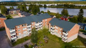 Flygfoto över två intilliggande flerbostadshus i tegel med gråa tak, omgivna av grönska. I denna fridfulla miljö av hyreslägenheter från Överkalixbostäder skapar en å och träd en fridfull bakgrund mot den klara himlen och framhäver det lugna bostadsområdet.