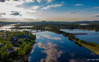 En naturskön flygfoto över en stad vid en flod vid solnedgången avslöjar hyreslägenheter inbäddade i det grönskande landskapet. Ån speglar himlen, med moln och blå nyanser som reflekteras på dess yta, när solljuset mjukt lyser upp Överkalixbostäder och en bro spänner över vattnet.