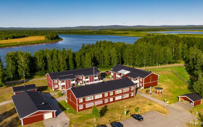 Flygfoto över röda träbyggnader, möjligen Överkalixbostäder hyreslägenheter, omgiven av täta gröna träd nära en stor, slingrande å under en klarblå himmel. Stigar och små vägar vävs genom denna fridfulla, naturliga miljö.
