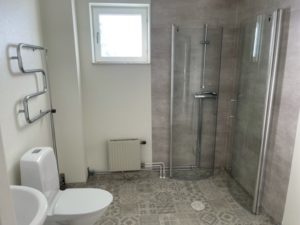 Ett modernt badrum i Överkalixbostäder har ljusgrå klinker på golv och väggar, en inglasad dusch, en väggmonterad värmare, en vit toalett, ett handfat, en handdukshängare och ett litet fönster som ger naturligt ljus. Perfekt för dig som söker bekväma hyreslägenheter.
