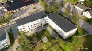 Flygfoto över ett bostadsområde med beiga hyreslägenheter och svarta tak. Det finns en grön gräsmatta med träd och en lekplats i mitten. En blå pil är ritad på trottoaren, som guidar dig mellan två byggnader. Foto Johan Olofsson.