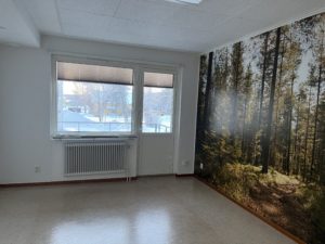 Ett tomt rum i Överkalixbostäder med ett stort fönster som visar snö utanför. En vägg har en väggmålning av en solbelyst skog, vilket skapar en inbjudande aura. Hyreslägenheterna har en radiator och ett vitt tak prydt med panelljus som ger värme och ljusstyrka.