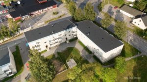 Flygfoto över ett bostadsområde med två stora, vita trevåningsbyggnader med platta tak omgivna av träd och gångvägar. En lekplats syns nedanför, och andra strukturer kantar gatan i bakgrunden, vilket framhäver Överkalixbostäders inbjudande hyreslägenheter.