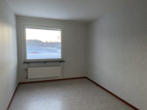 Ett tomt rum med ljusa väggar och golv i en av Överkalixbostäders hyreslägenheter. Ett stort fönster ramar in ett snölandskap och träd utanför, medan en radiator sitter under. Rummet är vackert upplyst med naturligt ljus.