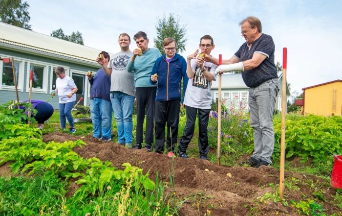 En grupp människor från Överkalixbostäder står tillsammans i en trädgård med växter och redskap. Vissa håller i skördade grönsaker, andra ler. En byggnad och träd, möjligen hyreslägenheter, syns i bakgrunden under en klar himmel.