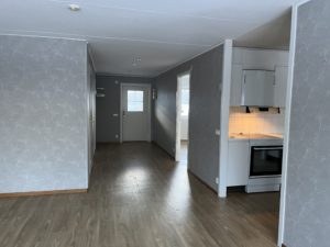 Ett tomt rum med grå väggar och trägolv, kännetecknande för Överkalixbostäders stil. En vit köksdel med ugn finns till höger. I bakgrunden släpper en dörr och fönster in naturligt ljus, perfekt för hyreslägenheter som söker ett mysigt men modernt utrymme.