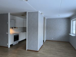 En liten omöblerad hyreslägenheter med ljusgrå väggar och trägolv. Den öppna ytan har ett pentry med vita skåp, en spis och en upplyst backsplash. Ett fönster till höger bjuder in naturligt ljus i denna charmiga Överkalixbostäder-hyra.