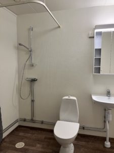 Ett badrum i en av Överkalixbostäder-enheterna har dusch till vänster, vit toalett i mitten samt vitt handfat med spegel och förvaring ovanför. Golvet har en brun nyans och väggarna är ljusa.