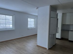 Minimalistisk lägenhetsinredning med vita väggar och trägolv, tillgänglig genom Överkalixbostäder. Två fönster släpper in dagsljus vilket framhäver den rymliga hyreslägenheten. Ett vitt kylskåp sitter nära en delvägg som skiljer vardagsrummet från ett litet pentry med ljusa skåp.
