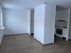 En minimalistisk lägenhetsinredning av Överkalixbostäder har en vardagsrumsdel med ljusa trägolv och vita väggar. Hyreslägenheterköket är delvis synligt, med vit spis och skåp. Naturligt ljus strömmar genom ett fönster till vänster.