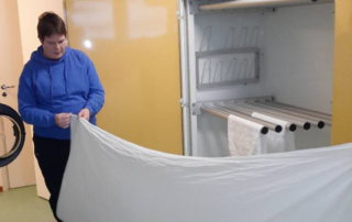 En person i en blå luvtröja håller ett lakan nära en stor torkmaskin med flera stavar, möjligen designad för att hänga och torka lakan. Lokalen, typisk för Överkalixbostäder hyreslägenheter, har ljusa väggar och stängd dörr.
