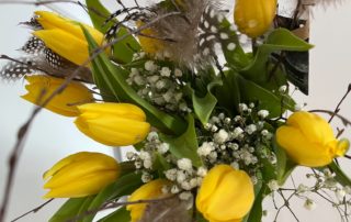 En bukett av livfulla gula tulpaner blandat med fina vita babyblommor framkallar Överkalixbostäders charm. Accenter av spräckliga fjädrar och gröna löv är varvade genomgående, vilket skapar ett livligt och elegant arrangemang mot en ljus bakgrund.