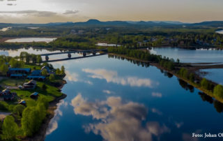 Flygfoto över ett fridfullt landskap nära Överkalixbostäder, med en flod som speglar den molniga himlen. Gröna träd och hus kantar flodstranden, där broar förbinder landet. Böljande kullar och en vidsträckt grönska ramar in denna charmiga hyreslägenheter i bakgrunden.