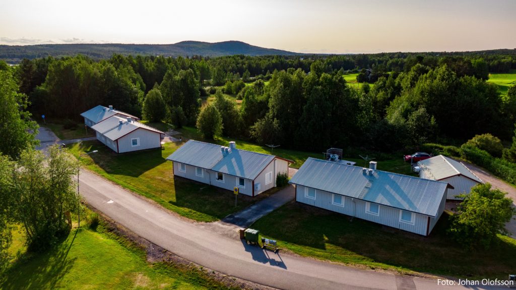 DJI_0731 text Flygfoto över en fridfull stadsdel Överkalixbostäder med enplanshus med plåttak, omgivna av grönskande träd. En väg slingrar sig genom detta lugna område av hyreslägenheter under en klar himmel, med avlägsna kullar som bidrar till det pittoreska landskapet.