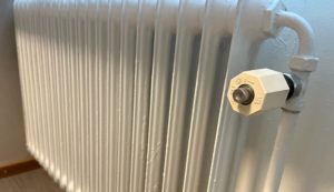 En vitmålad gjutjärnsradiator med sexkantsventil är monterad på en ljus vägg i Överkalixbostäder. En glimt av trägolv tittar igenom längst ner till vänster och förstärker den mysiga atmosfären som är typisk för hyreslägenheter.