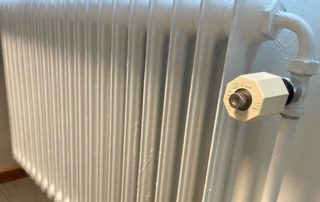 En vitmålad gjutjärnsradiator med sexkantsventil är monterad på en ljus vägg i Överkalixbostäder. En glimt av trägolv tittar igenom längst ner till vänster och förstärker den mysiga atmosfären som är typisk för hyreslägenheter.