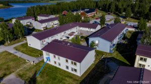 Flygfoto över ett bostadsområde med flera tvåvåningshus, troligen hyreslägenheter som förvaltas av Överkalixbostäder, inbäddat bland träd och gräs. En fridfull sjö pryder bakgrunden när en blå pil pekar på en byggnad. Himlen är klar och krispig.