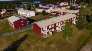 Flygfoto över ett bostadsområde med rött och beige Överkalixbostäder hyreslägenheter. Byggnaderna ligger inbäddade bland gräs och träd, med en smal väg som slingrar sig igenom. En blå pil markerar en byggnad nära floden, mot en bakgrund av fler strukturer.