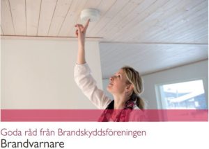 En person sträcker sig upp mot en rökdetektor monterad i taket i ett ljust rum, vilket speglar Överkalixbostäders engagemang för brandsäkerhet. Bilden innehåller svensk text med brandsäkerhetsråd från Svenska Brandskyddsföreningen, relevant för alla hyreslägenheter.