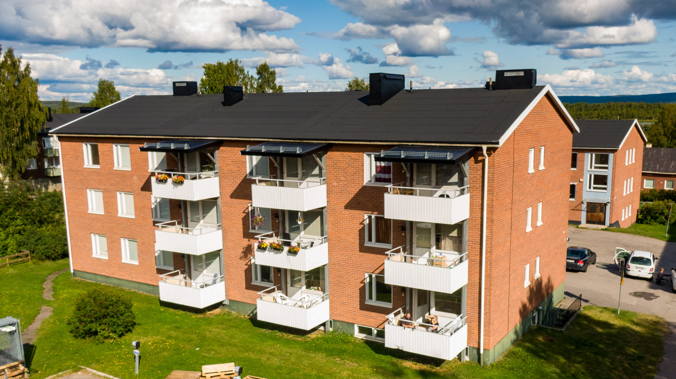 Detta charmiga bostad från Överkalixbostäder är ett trevåningshus i tegel med balkonger med blomkrukor, och har solpaneler på taket. Omgiven av gräs, träd och en halvmolnig himmel syns liknande hyreslägenheter i bakgrunden.