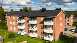 Ett hyreshus i rött tegel med tre våningar. Varje enhet har en vit balkong med en del med växter. Taket har solpaneler. Himlen är delvis molnig och träden omger byggnaden. Det finns en gräsyta framför.