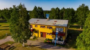 Flygfoto över ett charmigt gult tvåvåningshus ägt av Överkalixbostäder inbäddat bland lummiga träd och gräs. Huset ståtar med ett sluttande grått tak och röda detaljer. En blå pil pekar på en balkong på andra våningen, med en fridfull flod som rinner i bakgrunden under en klarblå himmel.