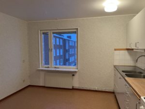 Ett litet tomt kök med beige golv och ljusa väggar. Den har ett vägghängt handfat med bänkskiva, vita skåp och ett fönster som visar utsikt över en tegelbyggnad utanför. En taklampa tänds.