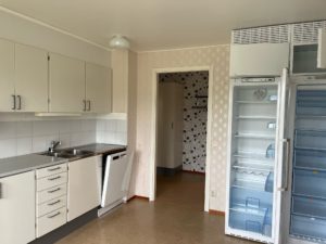 Kök med vita skåp, dubbelt handfat och diskmaskin. Till höger finns ett öppet, tomt kylskåp. Väggarna har mönstrade tapeter, och det finns en dörröppning som leder till ett annat rum. Golvet är beige.