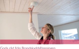 En kvinna sträcker sig upp för att testa en rökdetektor i ett trätak och ler när hon tittar på den. Texten nedan lyder, Goda råd från Brandskyddsföreningen, som påminner boende i Överkalixbostäders hyreslägenheter att hålla sig säkra.