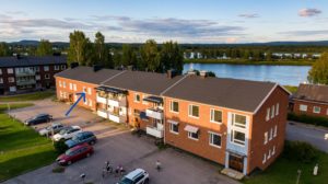 Flygfoto över ett bostadshus i tegel med flera balkonger. Flera bilar står parkerade på platsen framför, och några personer syns när de går eller cyklar. En flod och träd är i bakgrunden, med en molnig himmel ovanför.